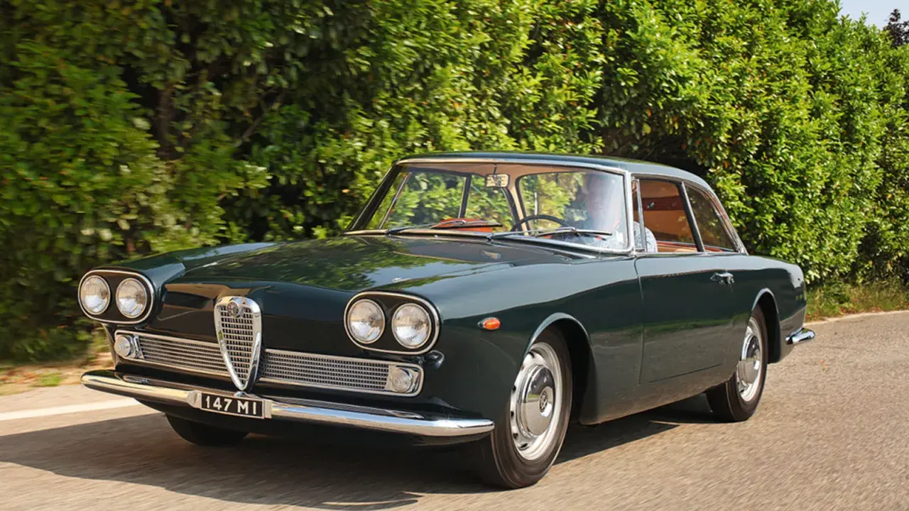 En mörkgrön Alfa Romeo Giulia Sprint GT kör längs en landsväg, en klassisk skönhet bland Alfa Romeo modeller.