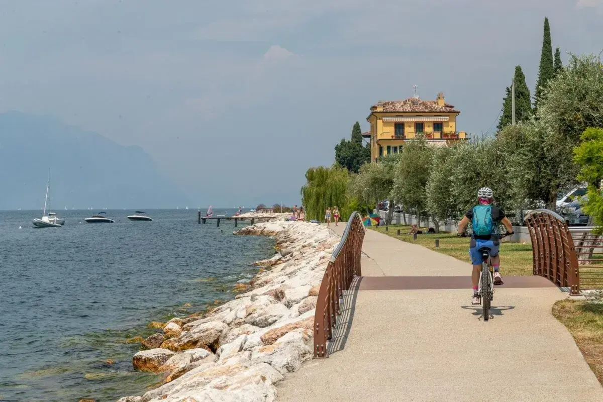 Ciclista sul lungolago, con barche e montagne sullo sfondo. Un assaggio di cosa vedere sul Monte Baldo, tra natura e relax.