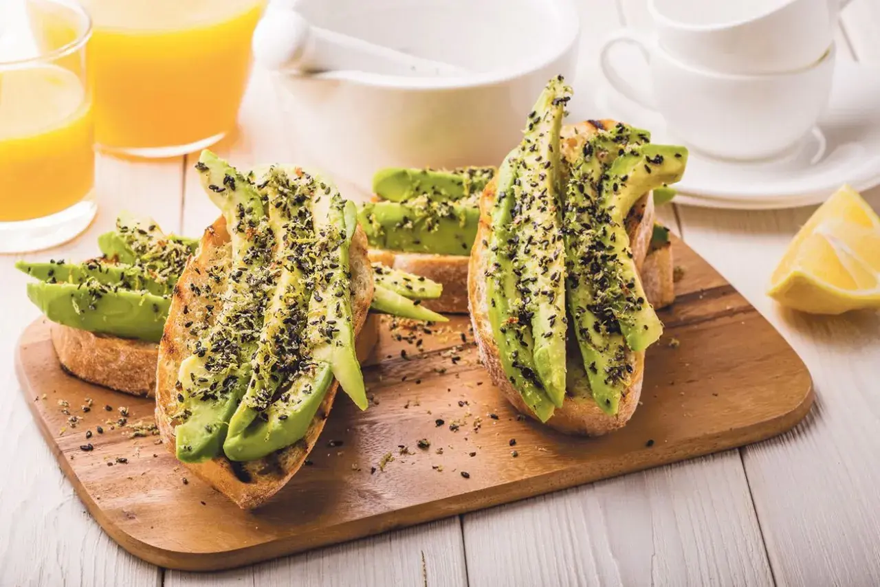 Tostadas con aguacate y semillas negras, perfectas para desayunos veganos. Acompañadas de zumo de naranja y limón.