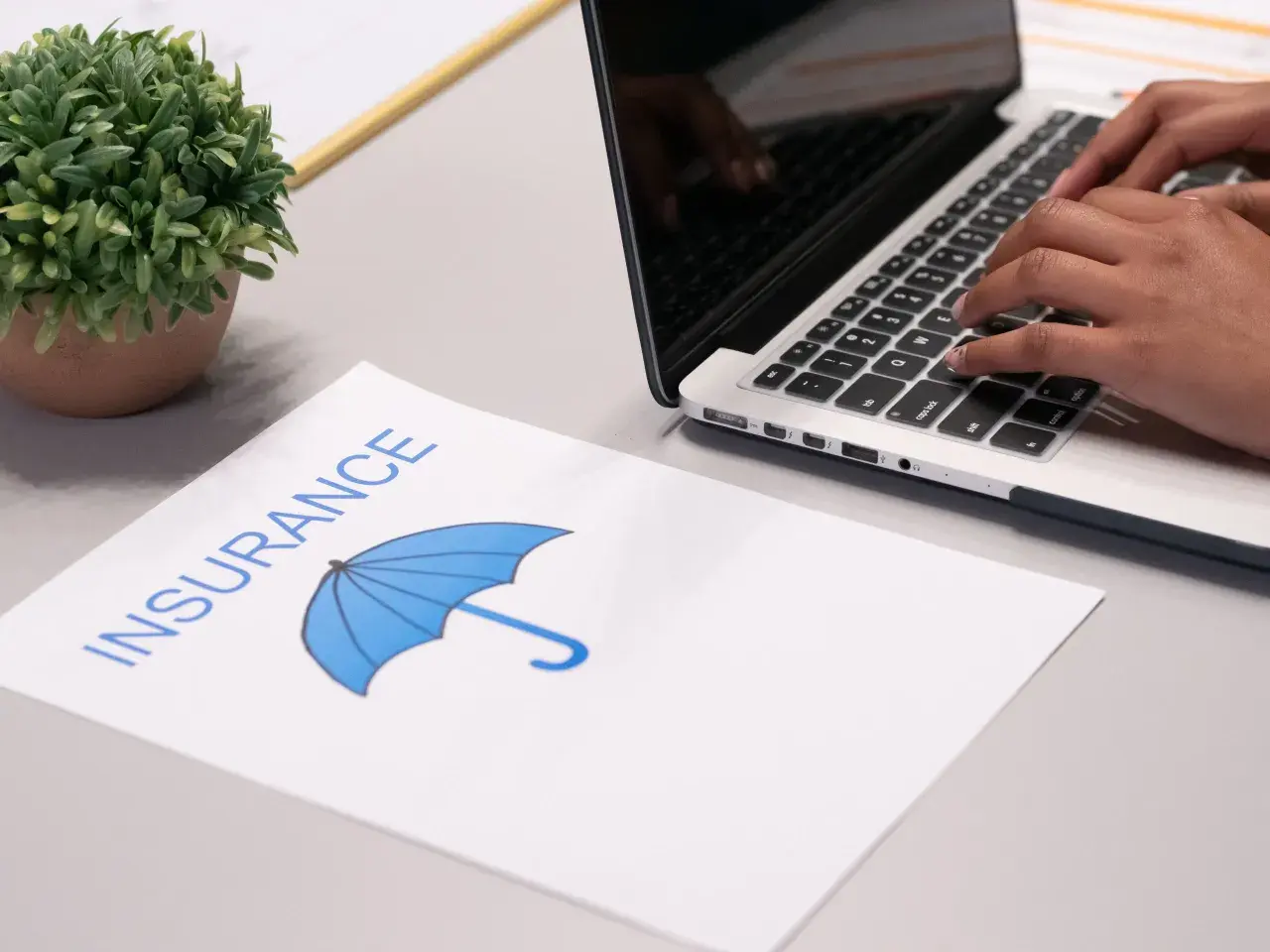Osoba wpisuje dane na laptopie, obok leży dokument z napisem "INSURANCE" i symbolem parasola, sugerujący ubezpieczenie domu od wszystkich ryzyk.