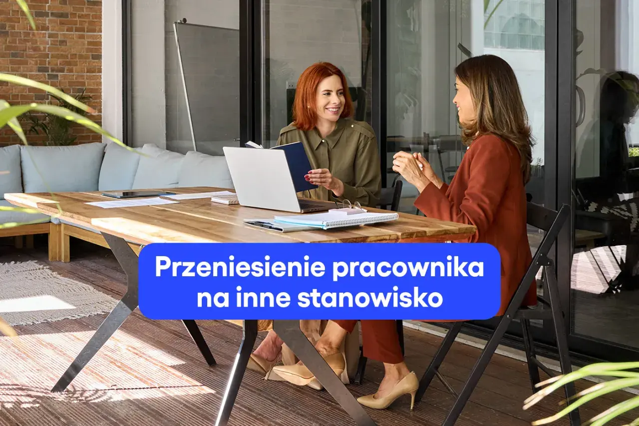 Dwie kobiety rozmawiają przy biurku, omawiając zmianę stanowiska pracy. Jedna z nich trzyma notatnik.