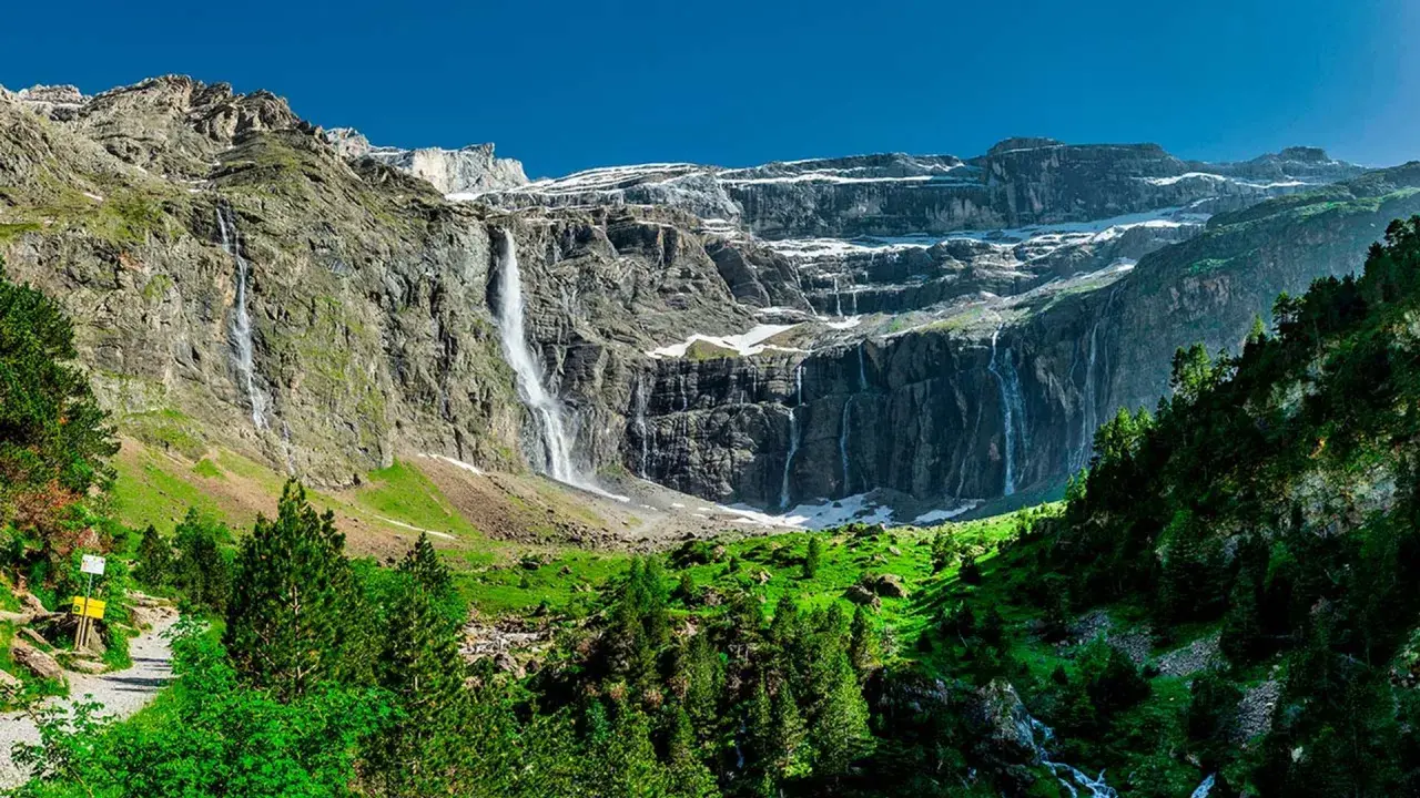 Cascadas impresionantes caen por acantilados rocosos cerca del pueblo de Gavarnie. Un sendero serpentea entre árboles verdes y prados exuberantes.
