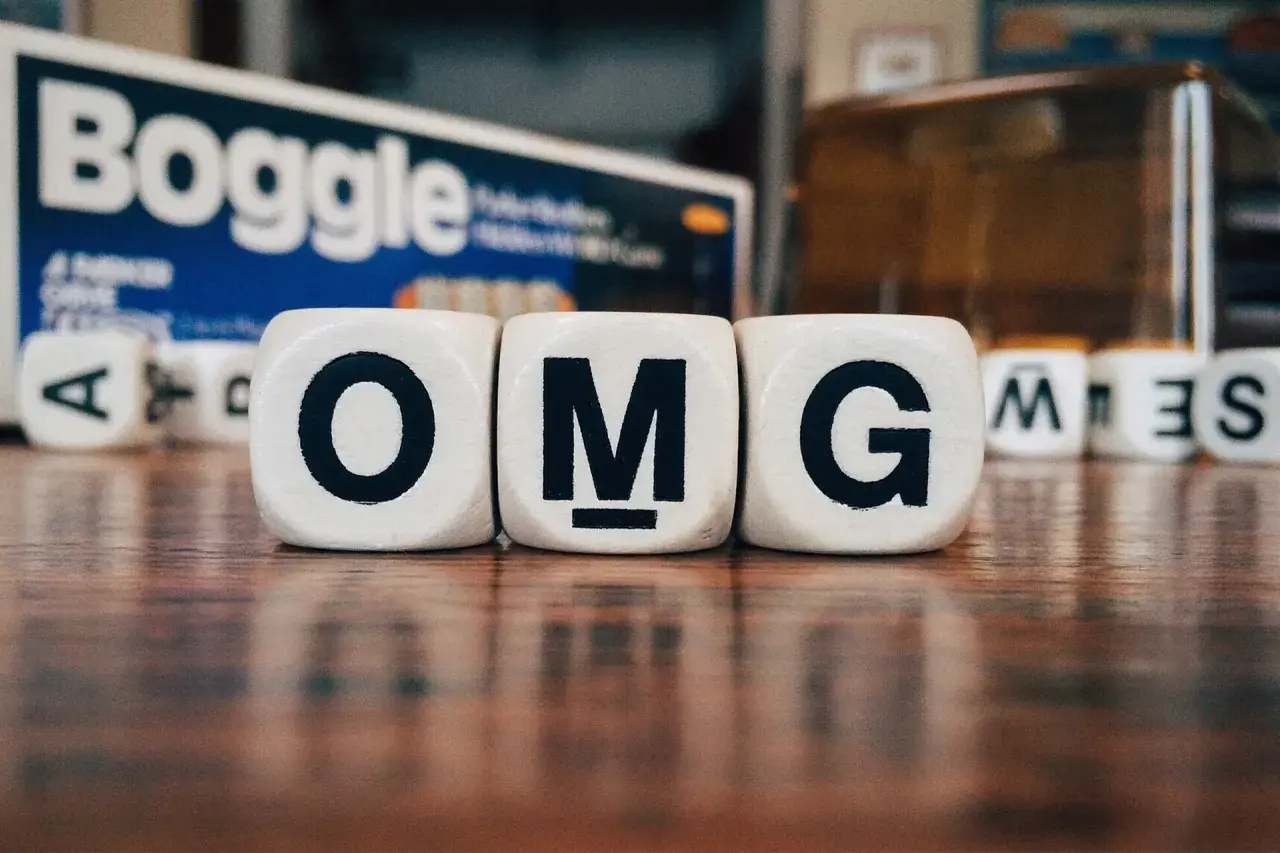 Dadi con lettere formano l'acronimo in inglese "OMG". Sullo sfondo, la scatola del gioco Boggle.