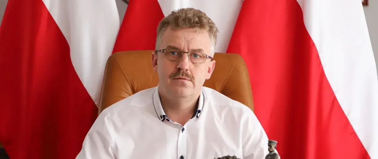 Starosta w okularach i białej koszuli, siedzący przed polskimi flagami, wygląda jakby właśnie skończył wykład o tym, co robi starosta na studiach.