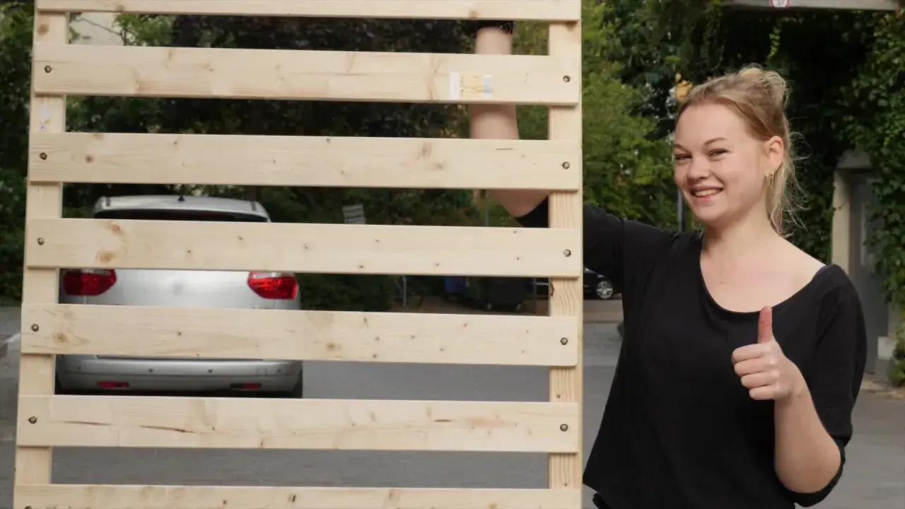 Junge Frau zeigt Daumen hoch neben selbstgebautem Lattenrost. Ein Auto ist im Hintergrund zu sehen.