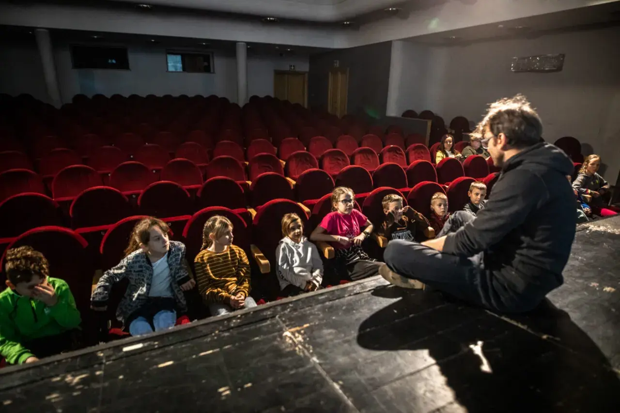 Dzieci słuchają opowieści na scenie, siedząc przed rzędem czerwonych foteli. To część repertuaru teatru Zagłębia.