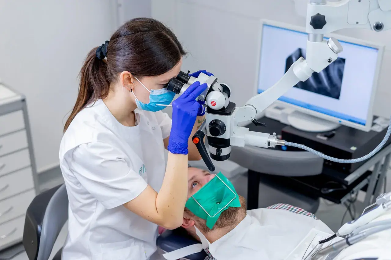 Dentystka w masce i rękawiczkach bada ząb pacjenta pod mikroskopem. Na ekranie widać zdjęcie rentgenowskie. To nowoczesne leczenie kanałowe, cena zabiegu zależy od skomplikowania.