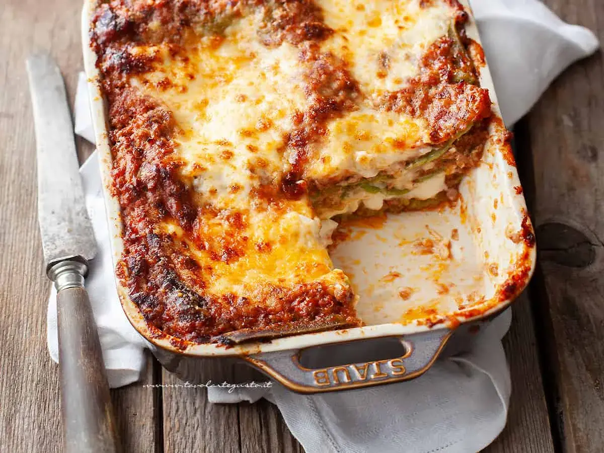 Lasagne alla bolognese appena sfornate, dorate e filanti, in una teglia Staub. Un coltello vintage è appoggiato accanto.