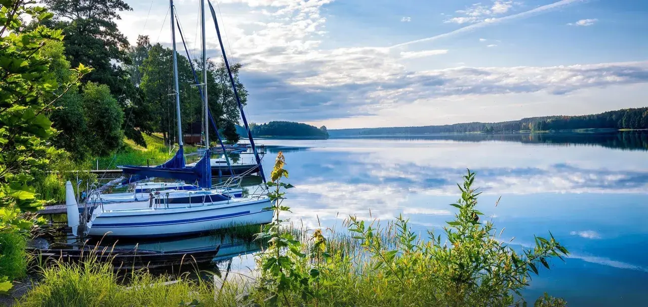 Żaglówki przy pomoście na spokojnym jeziorze. W tle las, a na wodzie odbijają się chmury. Mazury ciekawostki: idealne miejsce na relaks i żeglowanie.