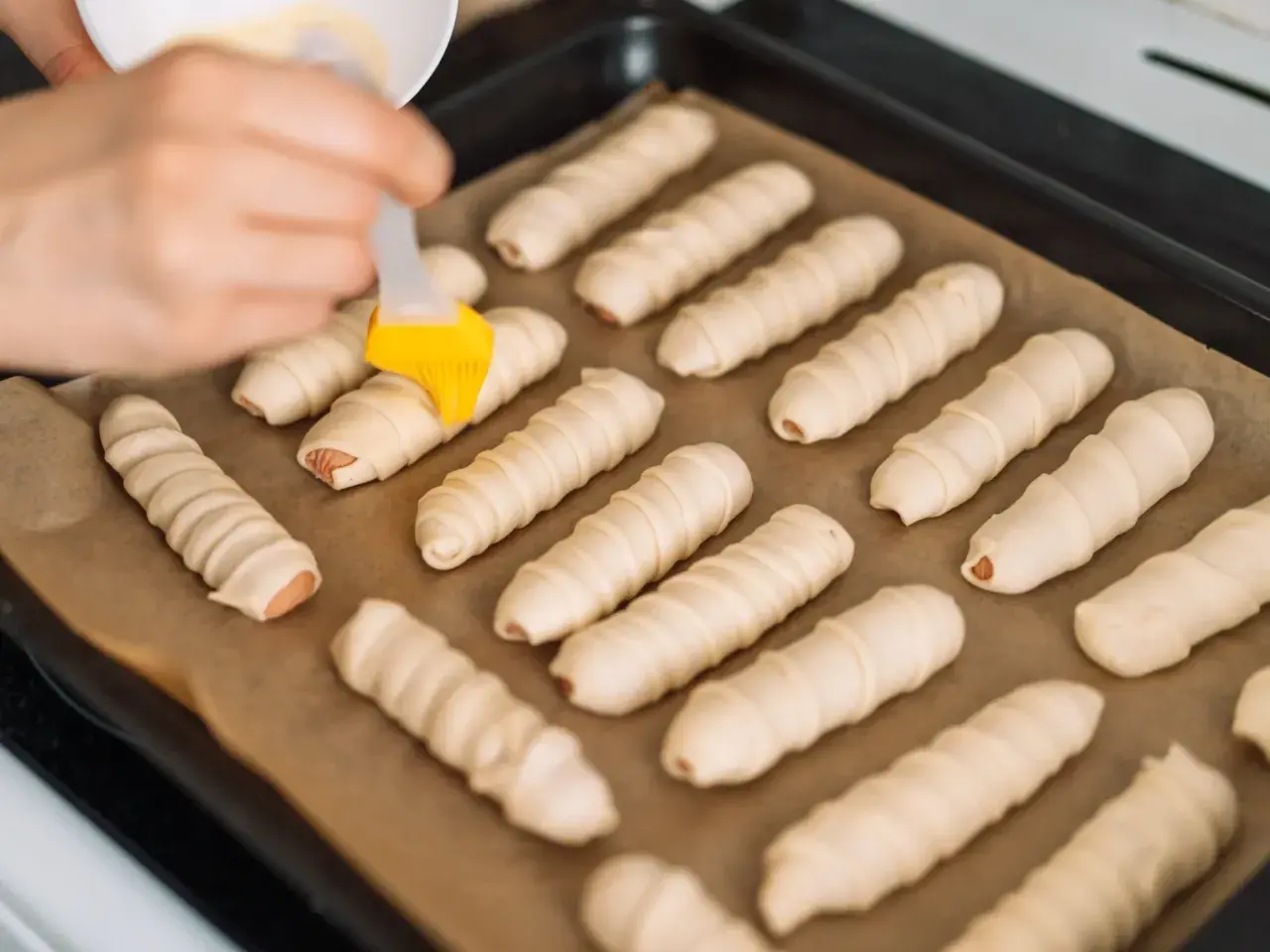 Ręka smaruje pędzelkiem parówki zawinięte w ciasto, przygotowując je do pieczenia. To prosty przepis na parowance.