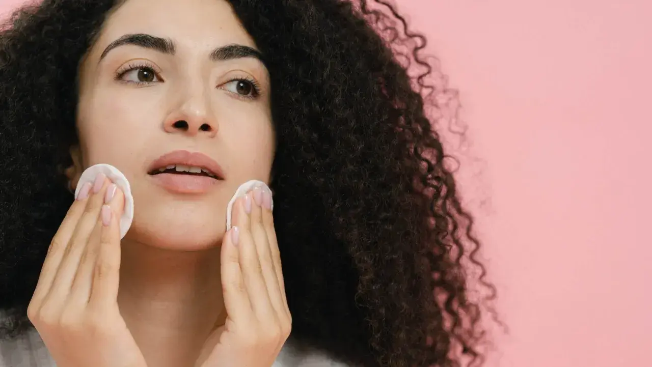 Donna con capelli ricci scuri pulisce il viso con dischetti di cotone, affrontando brufoli e punti neri per una pelle radiosa.