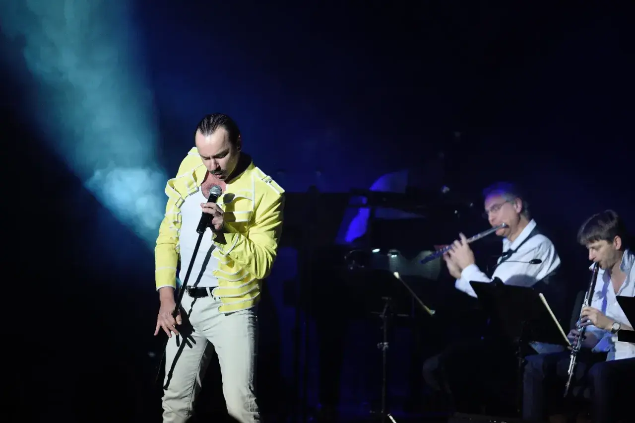 Wokalista w żółtej kurtce śpiewa na scenie, podczas gdy muzycy grają na instrumentach dętych. To koncert queen symfonicznie.