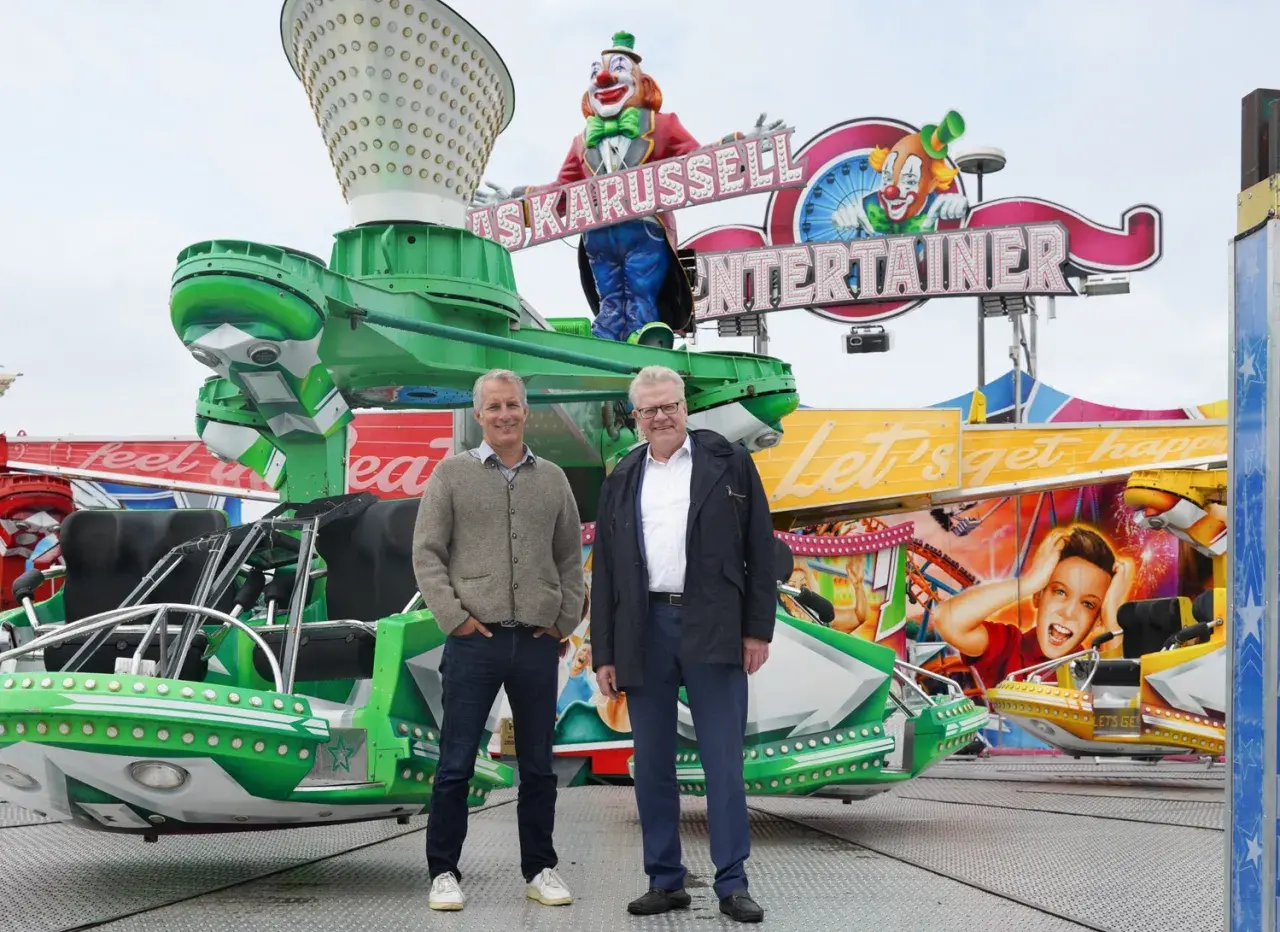 Zwei Männer stehen vor einer bunten Kirmesattraktion mit Clowns und der Aufschrift "Entertainer". Ein fröhlicher Ausblick auf das Bayreuth Herbstfest.