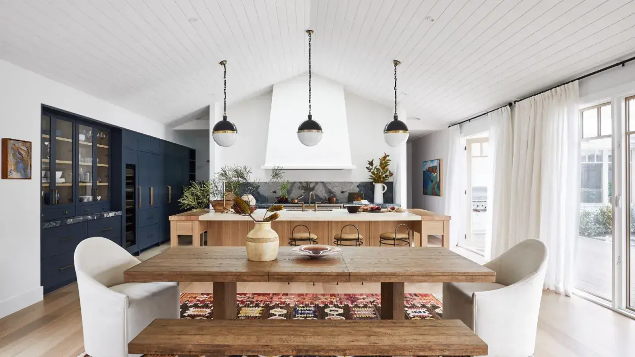 A modern lake house interior featuring a rustic wooden dining table, plush white chairs, and a sleek navy kitchen island.