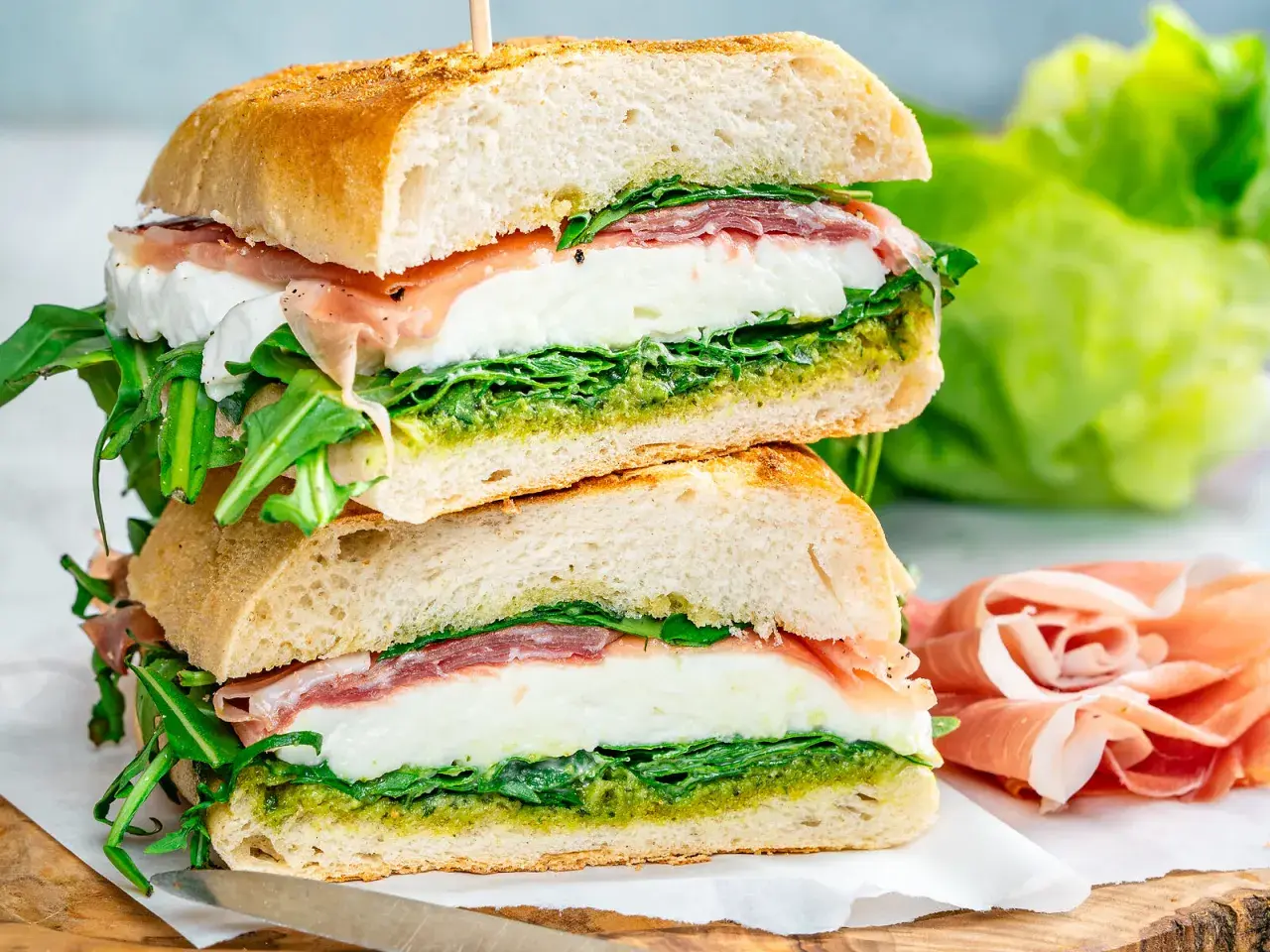 Dos bocadillos italianos apilados, rellenos de mozzarella fresca, prosciutto, rúcula y pesto, sobre pan rústico.