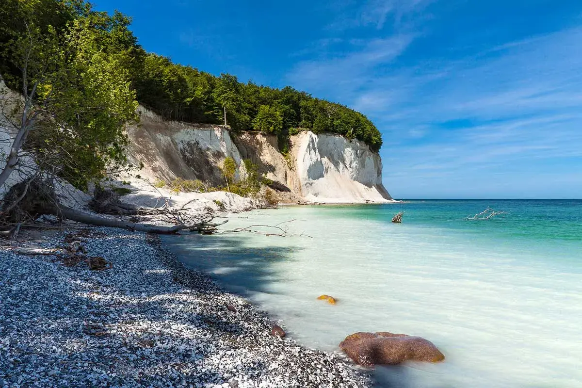 Weiße Kreidefelsen ragen steil auf, bewaldet von oben. Die Ostsee bei Sassnitz schimmert türkisblau, ein Kiesstrand lädt zum Verweilen ein.