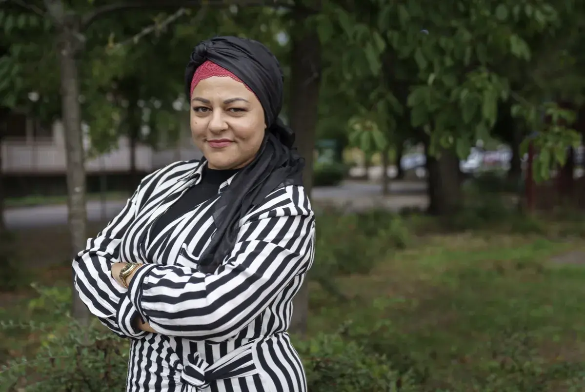 Zeina Mourtada, med en svart och röd turban och en randig jacka, står med armarna i kors utomhus.