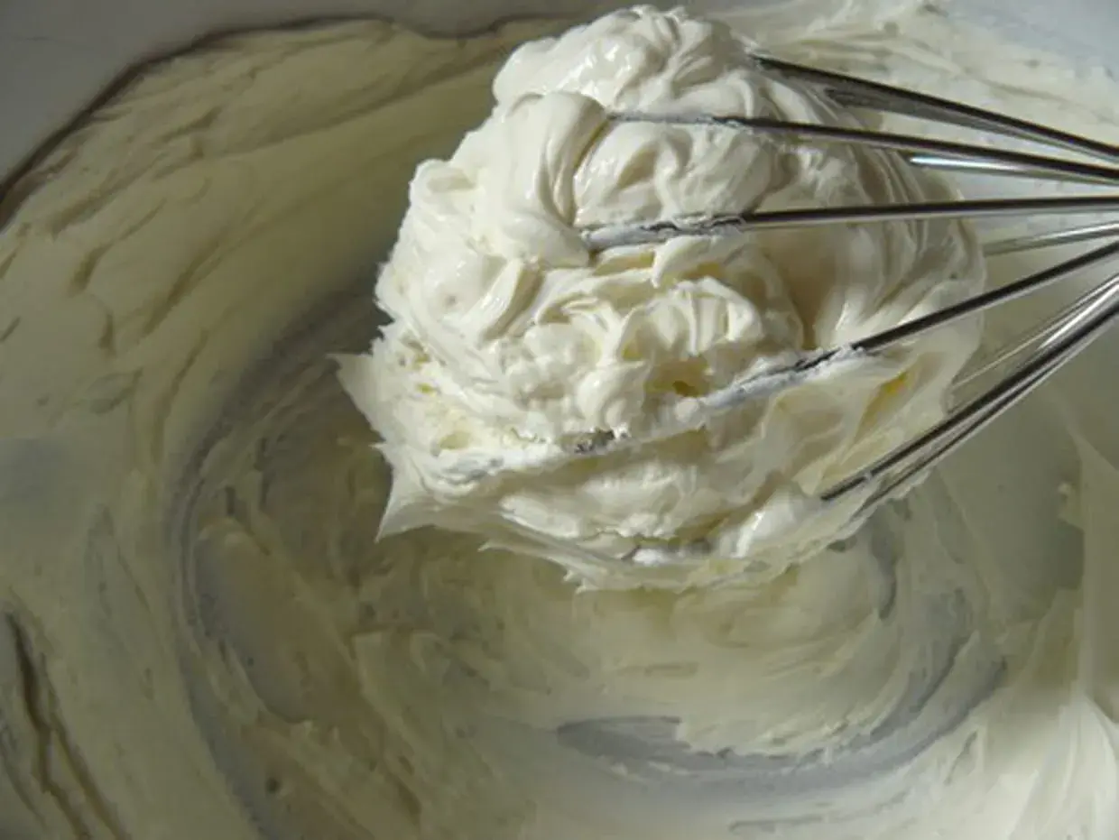 Un fouet de cuisine retient une généreuse portion de glacage mascarpone crémeux, prêt à garnir un gâteau.