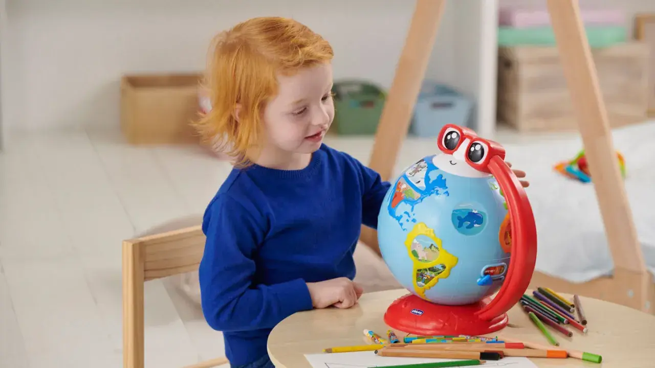 Un bambino di 5 anni con capelli rossi esplora il mondo con un globo interattivo, circondato da matite colorate.