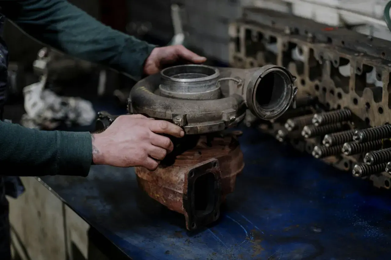 Mechanik trzyma turbosprężarkę, przygotowując się do jej wymiany krok po kroku. Obok leży głowica silnika.