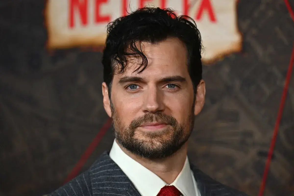 Henry Cavill w garniturze i czerwonym krawacie, z zarostem i niebieskimi oczami.
