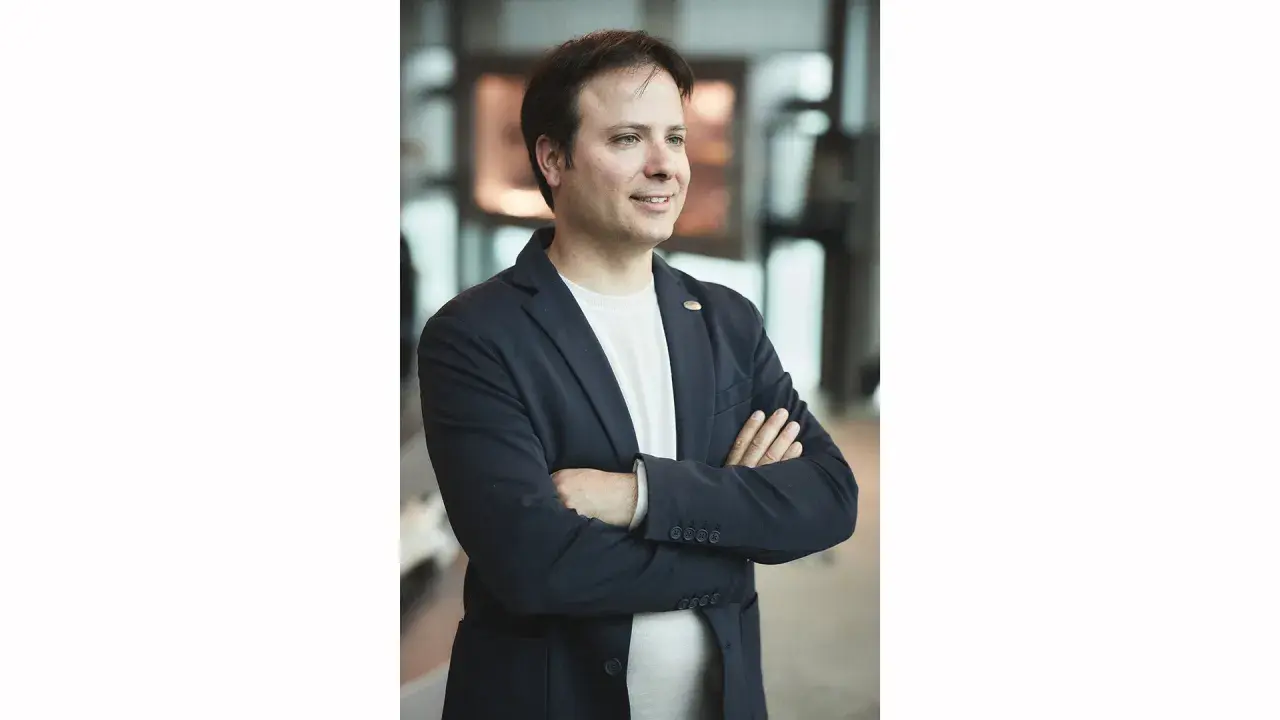 Christopher Pagani, mit verschränkten Armen und einem Lächeln, blickt nach rechts. Er trägt einen dunkelblauen Blazer über einem weißen T-Shirt.