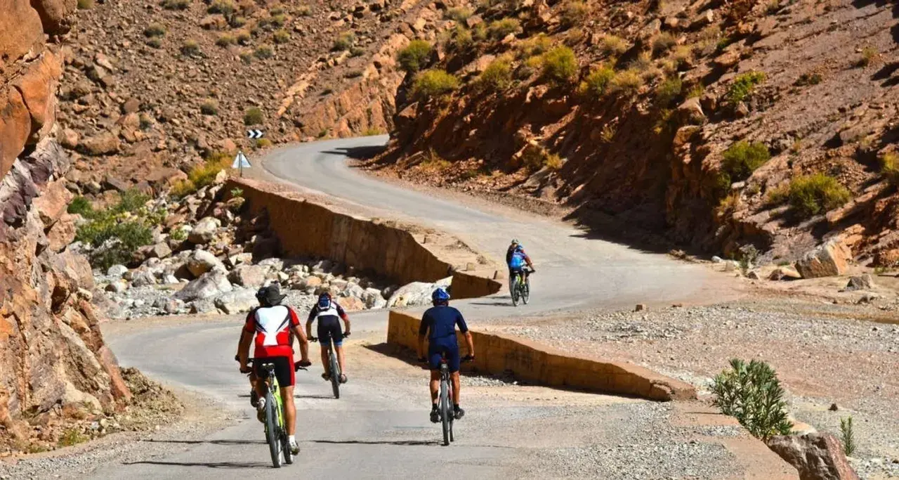 Ciclistas disfrutan de una aventura de **Marruecos en bici** por un camino sinuoso entre montañas rocosas y vegetación escasa.