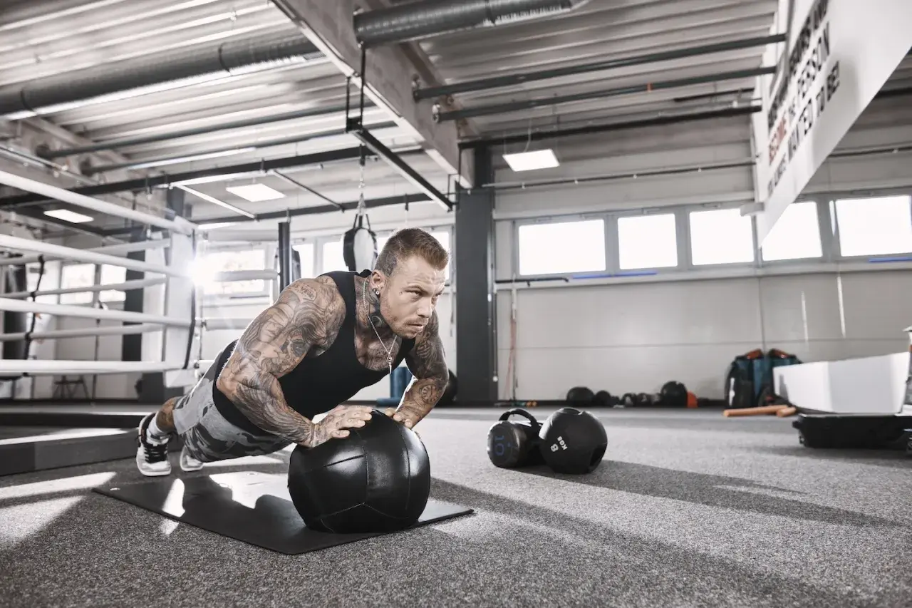 Mann mit Tattoos macht Liegestütze auf einem Medizinball, fokussiert auf sein hypertrophie training. Im Hintergrund ein Boxring und Trainingsgeräte.