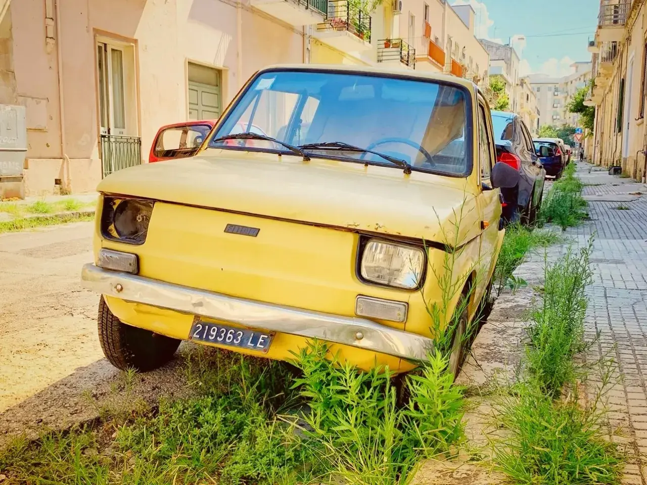 A vintage yellow Fiat, a symbol of driving in Puglia, sits parked on a sun-drenched street, overgrown with lush green grass.