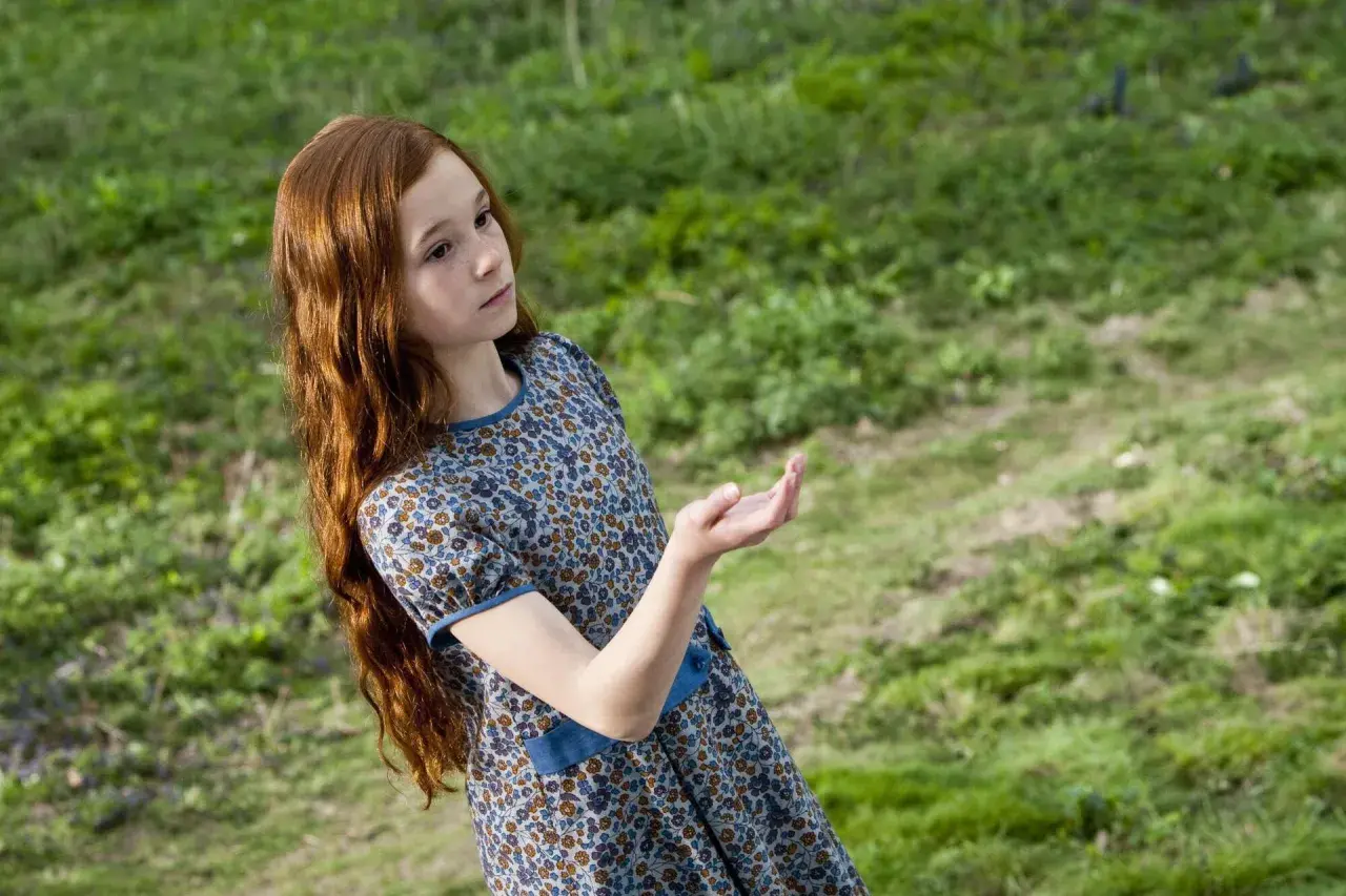 Ruda dziewczynka w kwiecistej sukience, Lily Potter, wyciąga dłoń w stronę zielonej łąki.