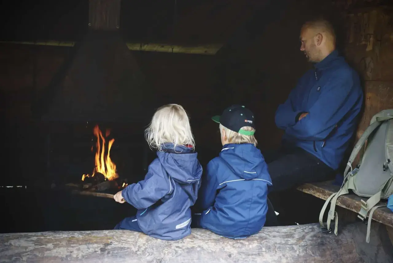 Två barn och en vuxen sitter vid en eld. En mysig paus under vandring med barn.