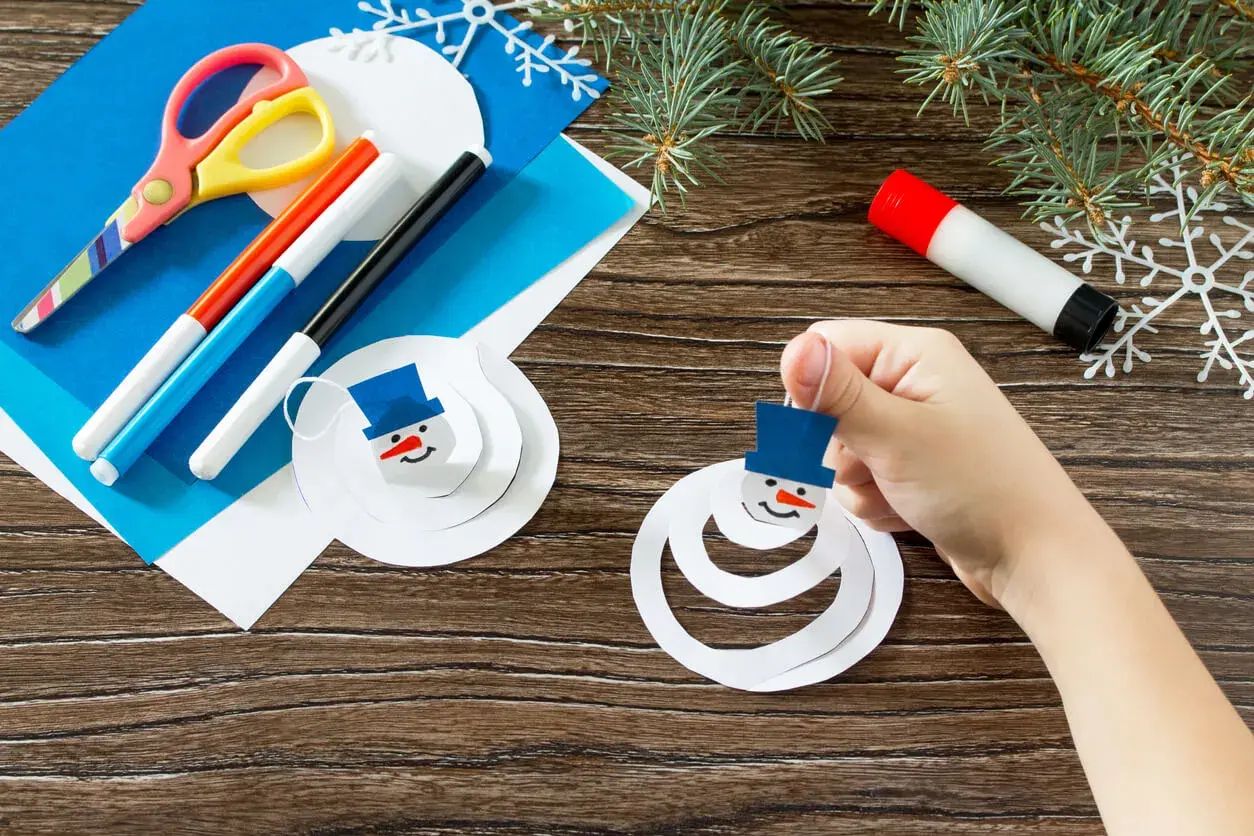 Niño crea muñecos de nieve con manualidades de invierno. Tijeras, rotuladores y pegamento listos para la diversión.