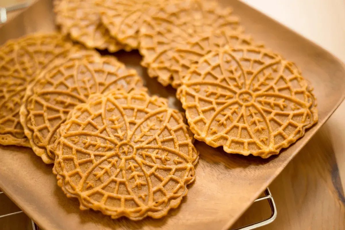 Pizzelle abruzzesi dorate e decorate con un motivo a fiore, disposte su un piatto di legno.