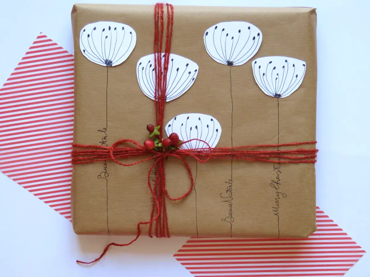 Confezioni pacchi regalo creativi con fiori stilizzati e bacche rosse, legati con spago rosso. Scritte "Buon Natale" e "Merry Christmas".