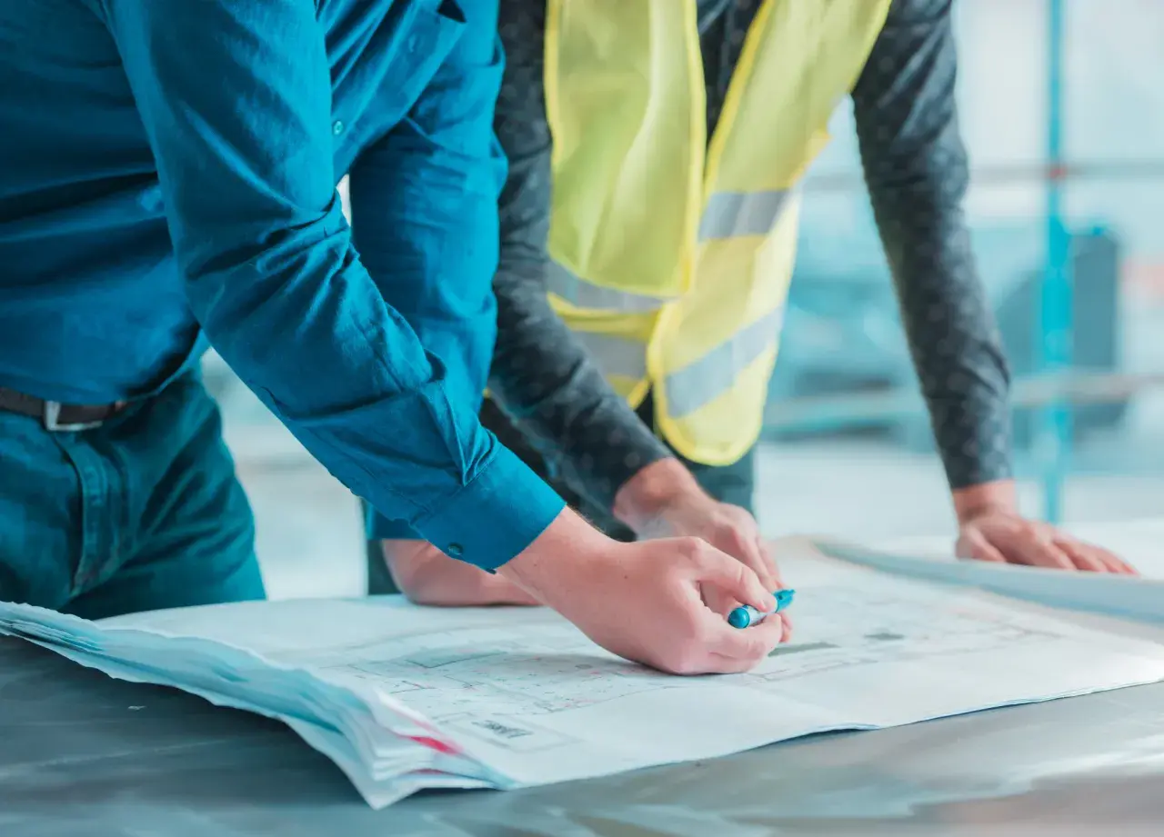 Deux hommes examinent des plans. L'un, en chemise bleue, pointe un détail avec un stylo. L'autre, en gilet jaune, pose ses mains sur les documents. Un constat huissier avant travaux est en cours.