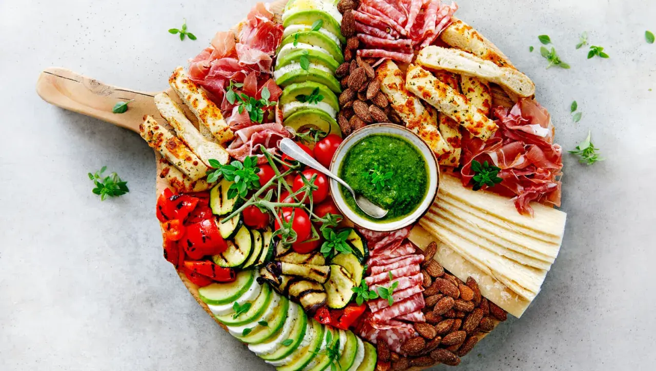 Tabla de antipasti italiani con prosciutto, salami, mozzarella, aguacate, tomates, pimientos y calabacín a la parrilla, acompañada de pesto y pan.