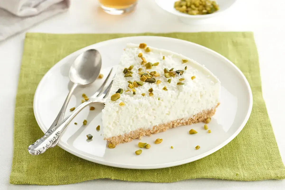 Rebanada de tarta de coco mousse, cubierta con coco rallado y pistachos picados, sobre plato blanco con cubiertos.