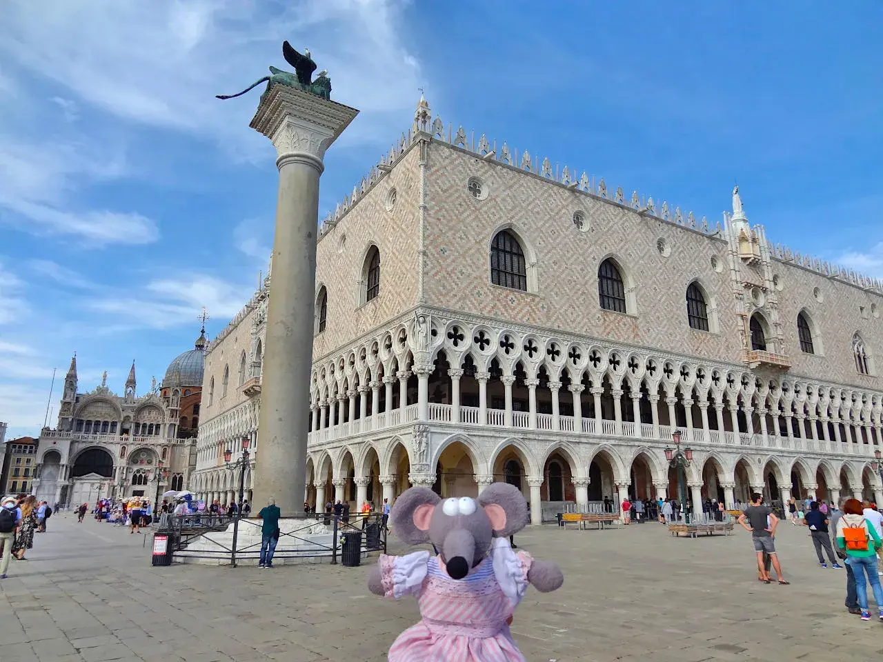 Une souris en robe rose devant l'entrée du Palais des Doges à Venise, avec la colonne de Saint-Marc et la basilique Saint-Marc en arrière-plan.
