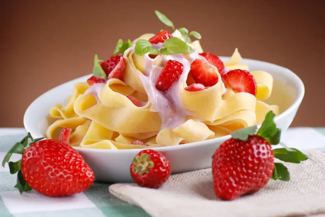 Pappardelle z kremowym sosem i kawałkami truskawek, ozdobione listkami mięty. Obok talerza leżą świeże truskawki.