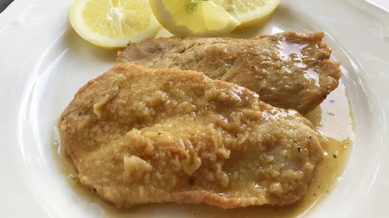 Deliziose scaloppine al limone, succulente e dorate, servite con spicchi di limone fresco su un piatto bianco.