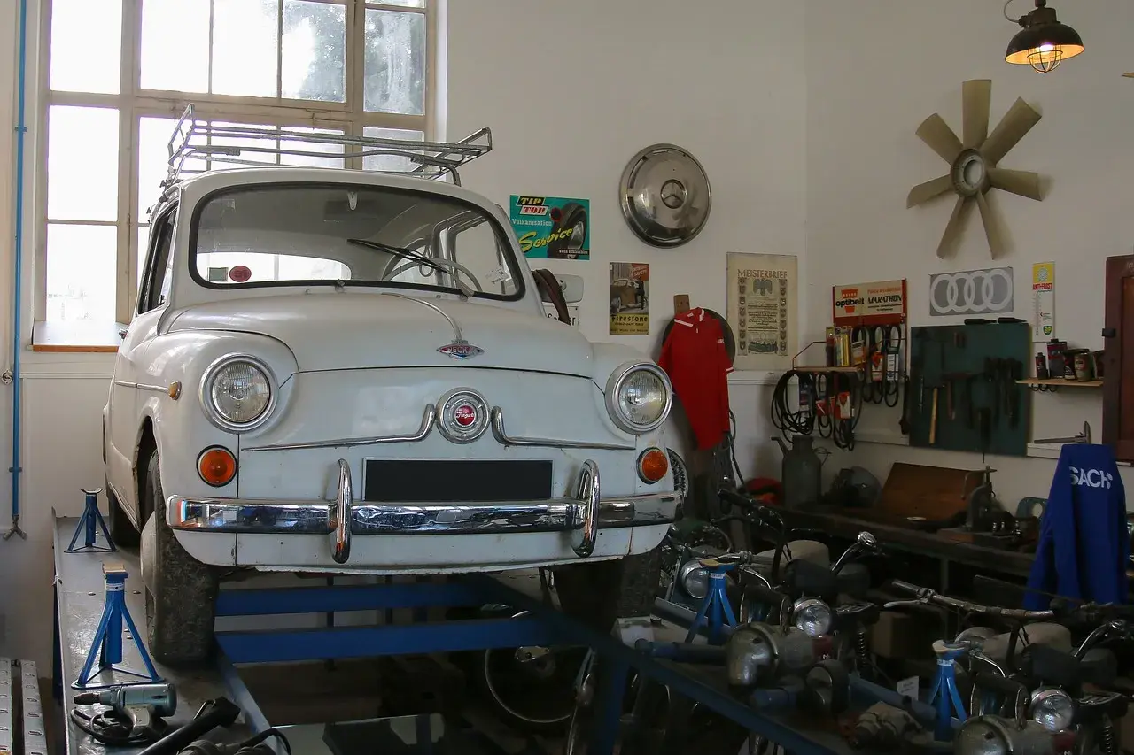 Biały Fiat 600 na podnośniku w warsztacie, gotowy do napraw. W tle narzędzia i motocykle, a nad nimi lampa zapewniająca oświetlenie kanału samochodowego.