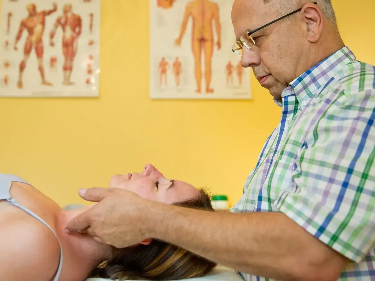 Ein Osteopath untersucht den Nacken einer Patientin. Anatomische Poster im Hintergrund illustrieren, was Osteopathie ist: eine ganzheitliche manuelle Therapie.