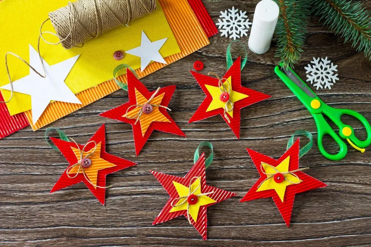 Kolorowe gwiazdki świąteczne z papieru, gotowe do zawieszenia. W tle materiały do ich wykonania: sznurek, nożyczki, klej.