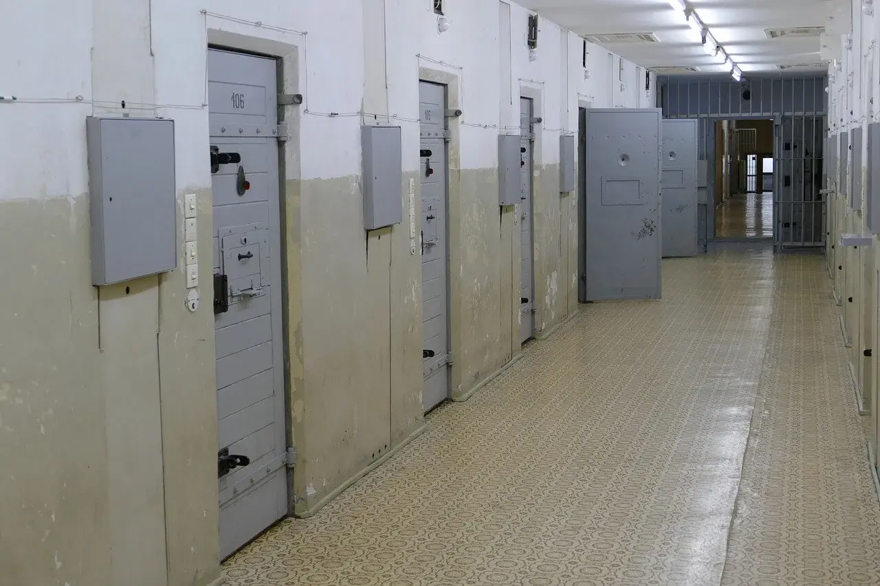 Korytarz więzienny z celami po obu stronach. Na drzwiach jednej z cel widnieje numer 106. W tle kraty i kolejne drzwi.