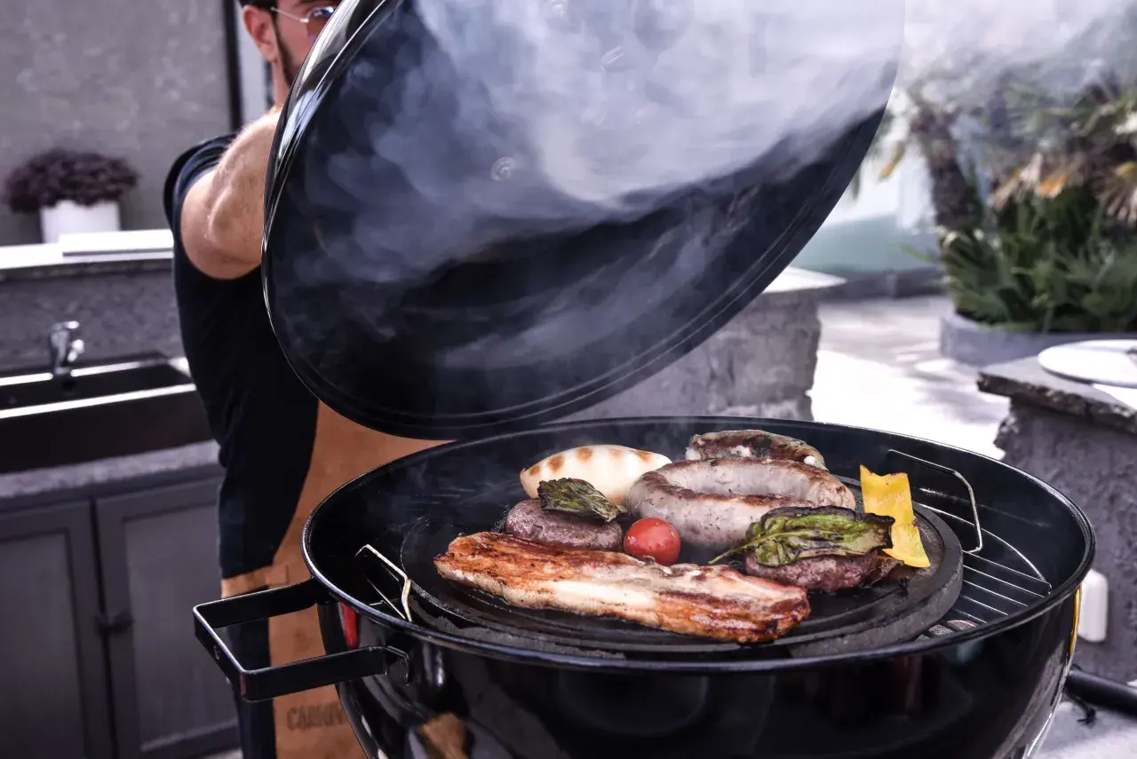 Un homme soulève le couvercle d'un barbecue fumant, révélant une sélection alléchante de viande au barbecue : saucisses, steaks, lard et légumes grillés.