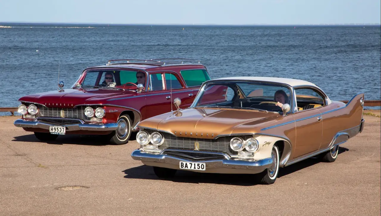 Två klassiska Plymouth Fury 1960, en röd kombi och en guldfärgad coupé, står vid havet.