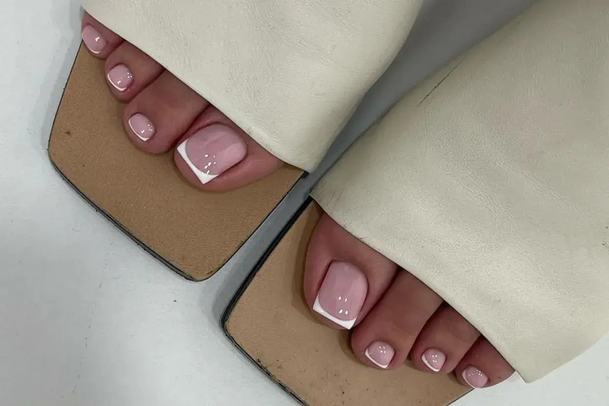 Elegancki pedicure french na różowych paznokciach stóp, ozdobionych białymi końcówkami i kropkami, w beżowych klapkach.