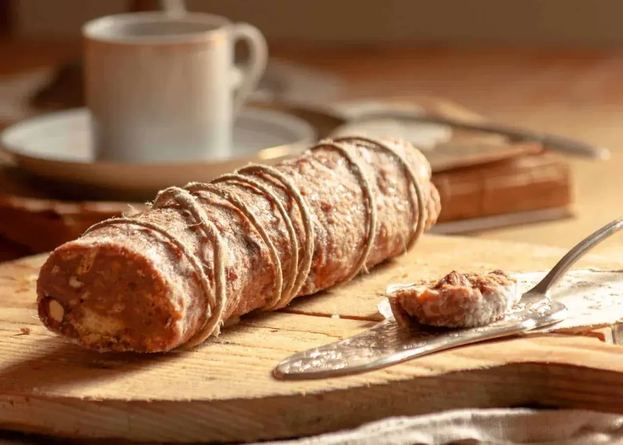 Ein rustikaler salame al cioccolato, gebunden mit Juteschnur, liegt auf einem Holzbrett. Daneben eine Kaffeetasse und ein Stück auf einem Tortenheber.