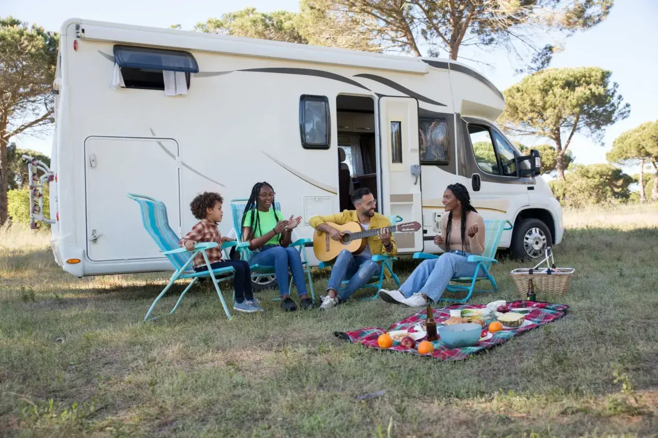 Familie genießt das Zelten mit Kindern. Ein Mann spielt Gitarre, während die anderen singen und lachen. Ein Picknickkorb und Obst liegen auf einer Decke.