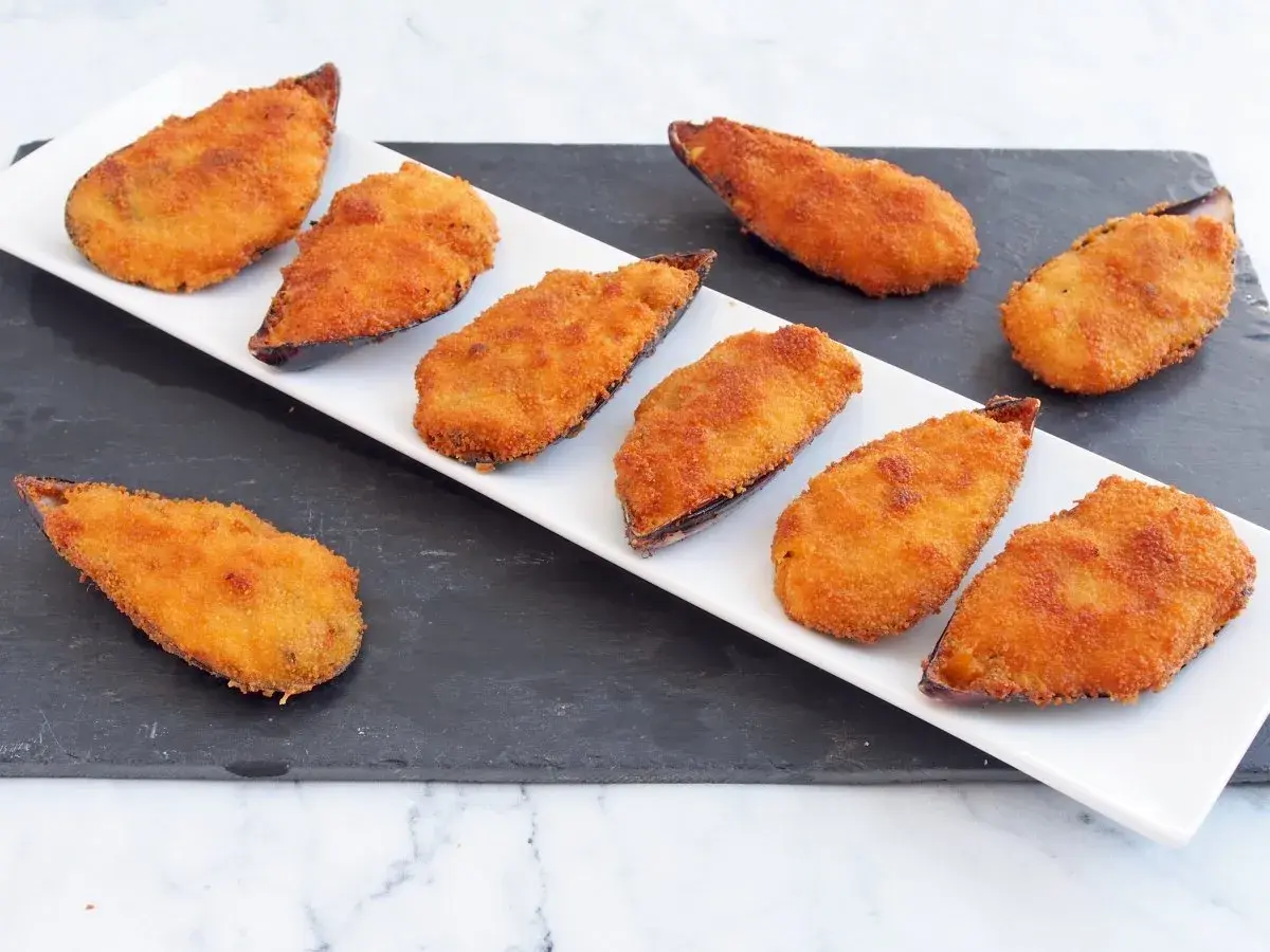 Ocho mejillones tigre dorados y crujientes, servidos en una bandeja blanca sobre una pizarra.